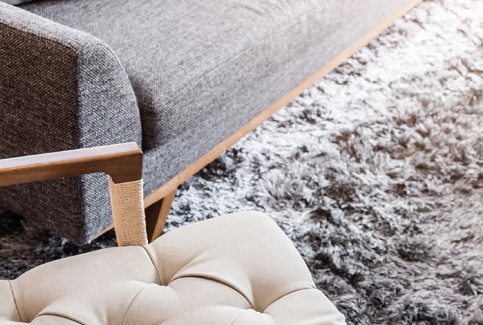 Detailaufnahme eines weichen Wohnzimmerteppichs neben Sofa und gepolstertem Sessel mit Holzarmlehne. Beispiel für gemütliche Wohnraumgestaltung mit Teppich und modernen Polstermöbeln